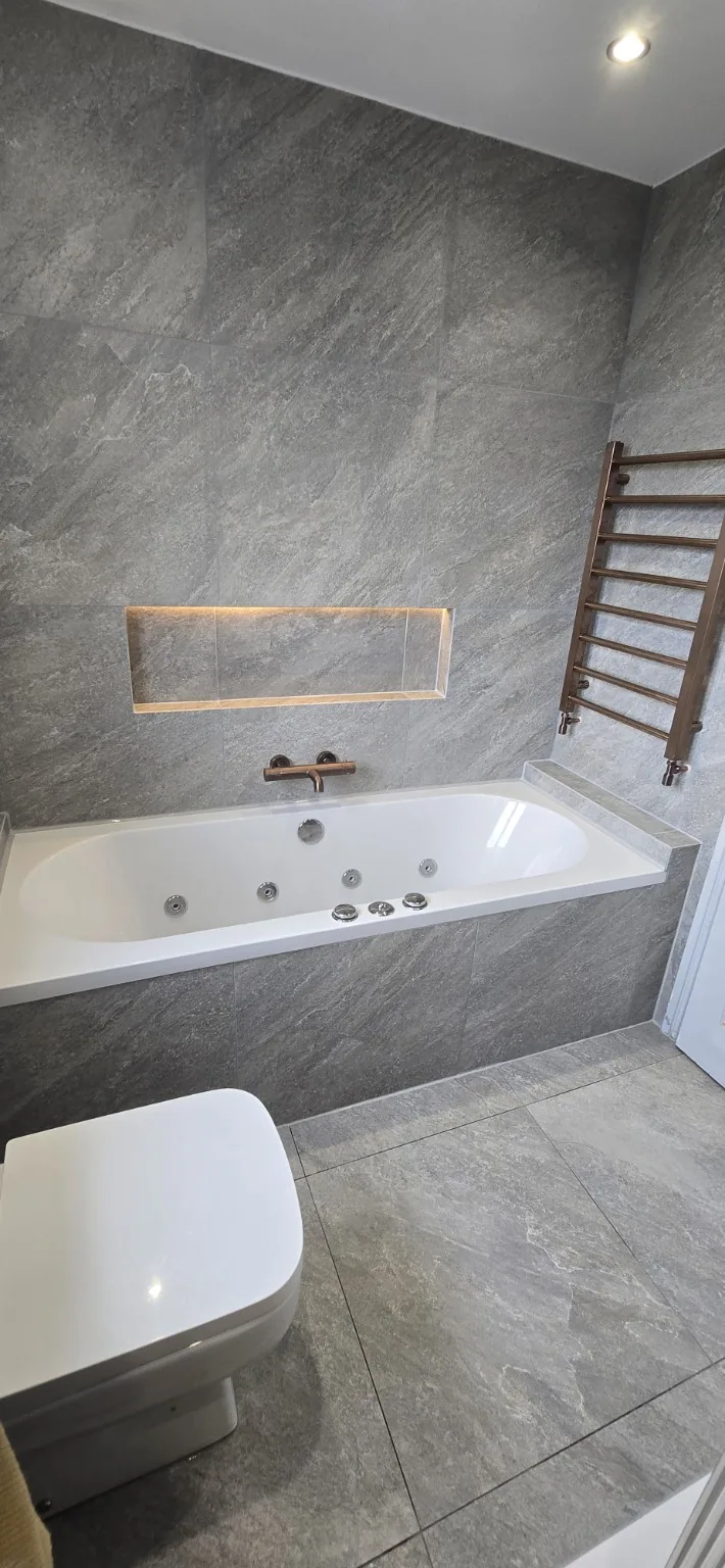 Completed Failsworth bathroom with a jetted bath, illuminated wall niche and grey wall and floor tiles.