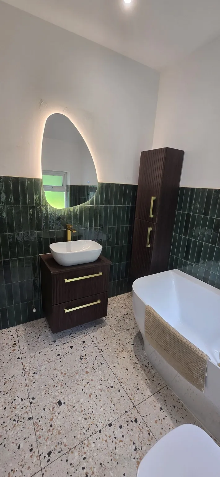 New basin and dark vanity cabinet beneath an illuminated mirror, with green wall tiles at the Worsley bathroom.